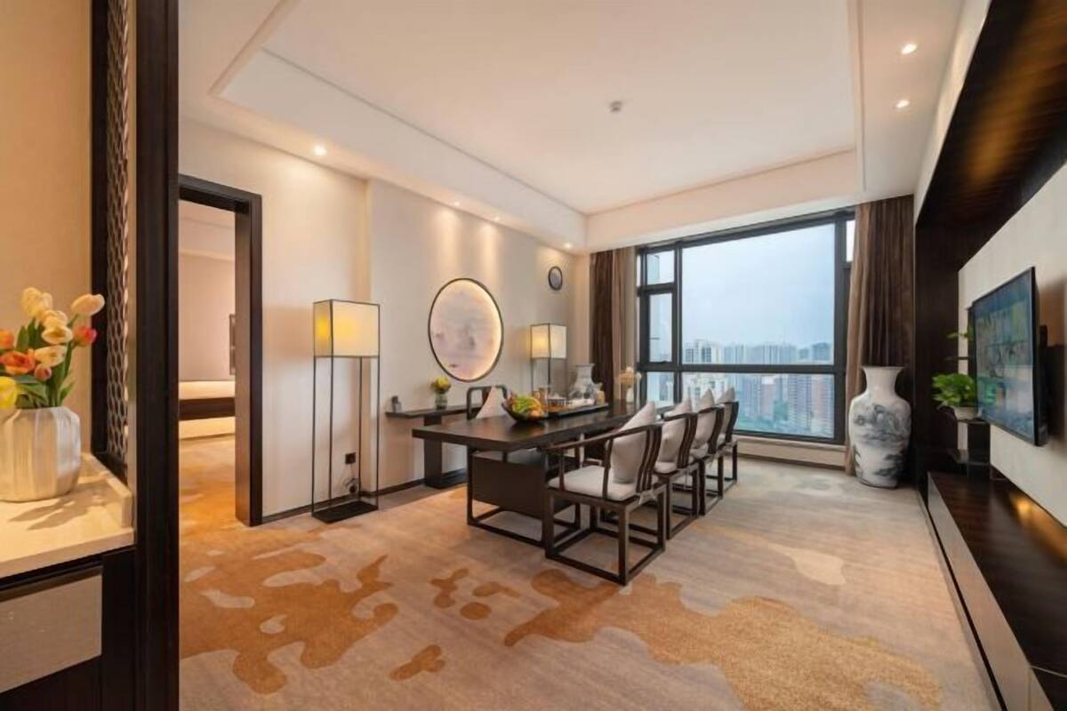 A modern hotel room with a large window, a dining area with a table and chairs, and a decorative vase on the side table.