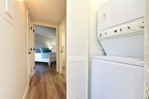 Un pasillo estrecho con una lavadora y secadora blancos, una cama con almohada azul y una puerta que da a un dormitorio.
