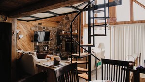 Ein gemütliches Blockhaus-Interieur mit Steinwand, Holzbalkendecke, Fernseher, Kamin und Wendeltreppe.