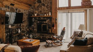Ein gemütliches Blockhäuschen-Interieur mit Kamin aus Stein, Holzwänden, einem Fernseher und bequemen Sitzgelegenheiten.