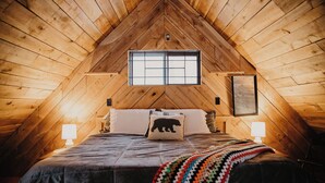 Ein gemütliches Schlafzimmer im Dachgeschoss mit Holzbalkendecke, einem großen Bett mit bunter Decke, zwei Nachttischlampen und einem Fenster mit Blick.