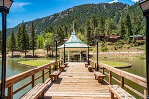 A wooden pier leading to a gazebo with benches, surrounded by a lake and forested mountains.