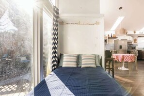 A bedroom with a large window, a bed with striped pillows, a dining area with a table and chairs, and a kitchen in the background. - Aptos Beach Retreat Hot Tub & 5 Min Walk to Sand (Aptos)