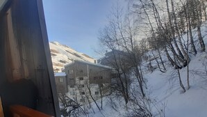 Näkymä ikkunasta, jossa on puinen kaide. Ikkunasta avautuu näkymä rakennukseen, jossa on useita ikkunoita, sekä lumipeitteiseen maisemaan, jossa on paljaita puita.