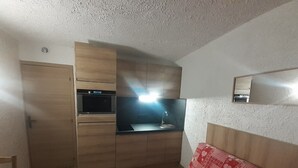Kompakti keittiö, jossa on mikroaaltouuni, uuni ja pesuallas. Siellä on puinen ovi ja pieni pöytä. Seinää vasten on punakuvioinen sohva.