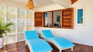 A sunlit room with two lounge chairs, a wooden ceiling, a white wall, and a window with wooden shutters.