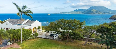 Ein Küstengrundstück mit einem weißen Gebäude, einer Rasenfläche und Blick auf das Meer und ein Bergmassiv.