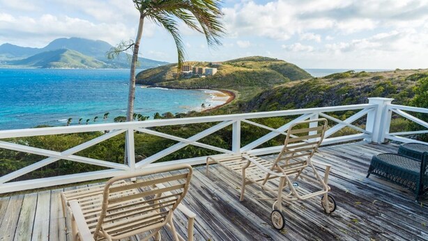 Eine Holzterrasse mit zwei Korbstühlen, mit Blick auf einen Strand und einen Berg im Hintergrund.