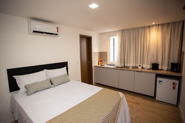 A hotel room with a bed, a small kitchenette, a window with curtains, and an air conditioning unit.