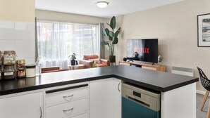 A modern kitchen with a black countertop, white cabinets, and a dishwasher. There is a large window with sheer curtains, a plant, and a sofa in the background. A flat-screen TV is mounted on the wall displaying the Netflix logo. - Modern Luxurious Living With Your Own Parking Spot (Sandy Bay)