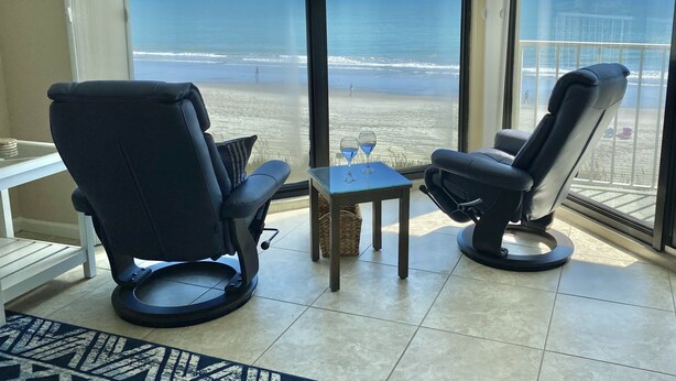 A room with two black leather chairs facing a large window overlooking a beach. A small table with two wine glasses is in front of the chairs. - Luxury Ocean Front (Murrells Inlet)