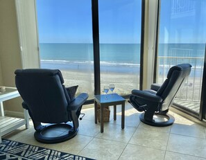 Una stanza con due poltrone in pelle nera rivolte verso una grande finestra con vista sulla spiaggia. Un tavolino con due bicchieri da vino è posizionato di fronte alle poltrone.