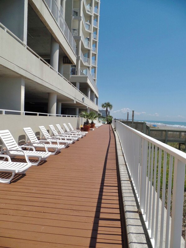 A sun deck with white lounge chairs and a wooden deck, overlooking a beach and a building with balconies. - Luxury Ocean Front-3rd floor (Murrells Inlet)