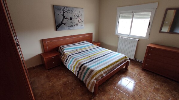 Une chambre à coucher comprenant un lit, des tables de chevet, une commode, un radiateur et une fenêtre avec des stores.