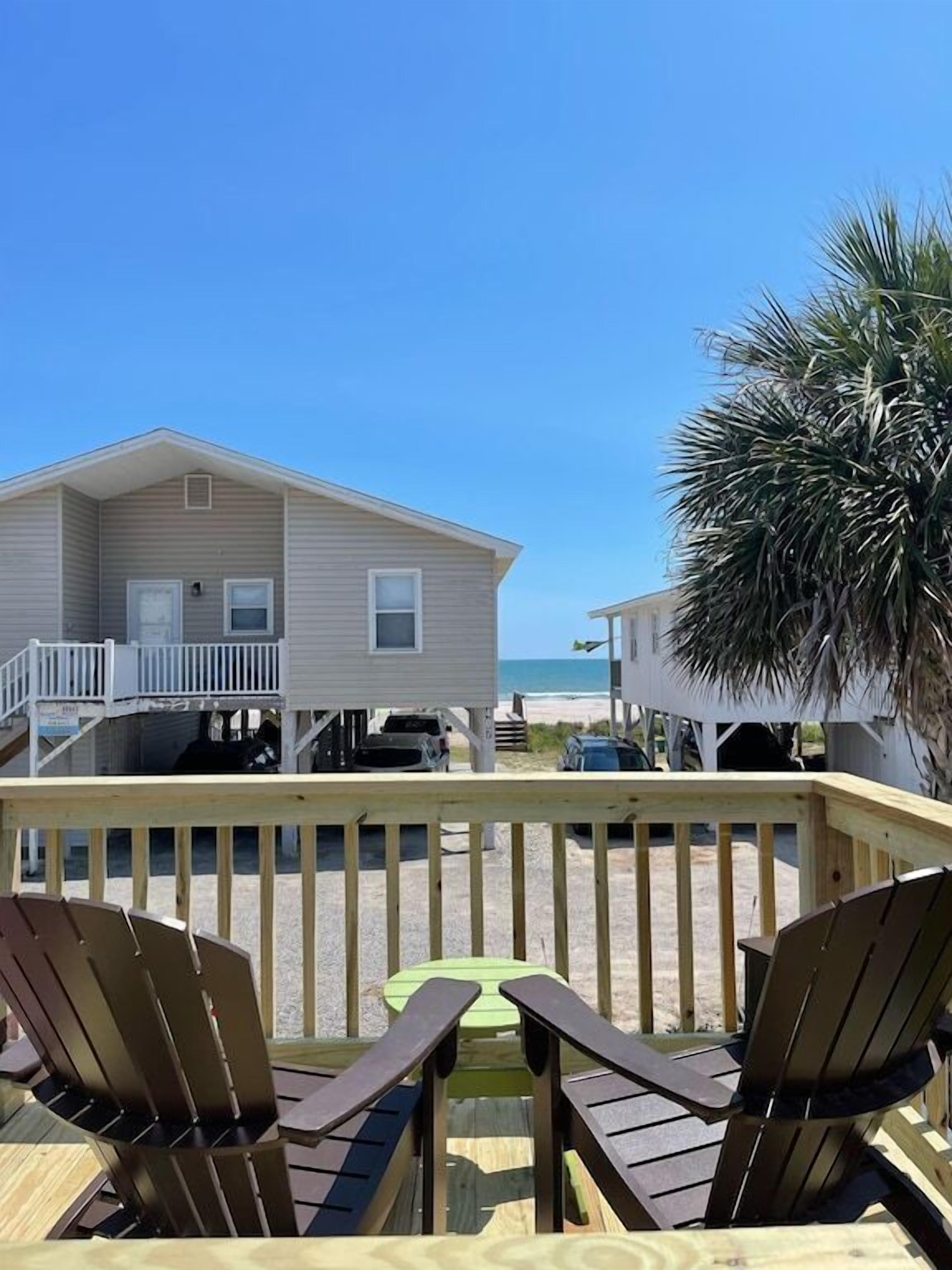 A beach house with a balcony, two Adirondack chairs, and a view of the ocean.