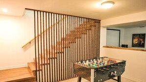 Un escalier avec des marches et une rampe en bois, menant à un palier où se trouvent une table de baby-foot et un tableau encadré accroché au mur.