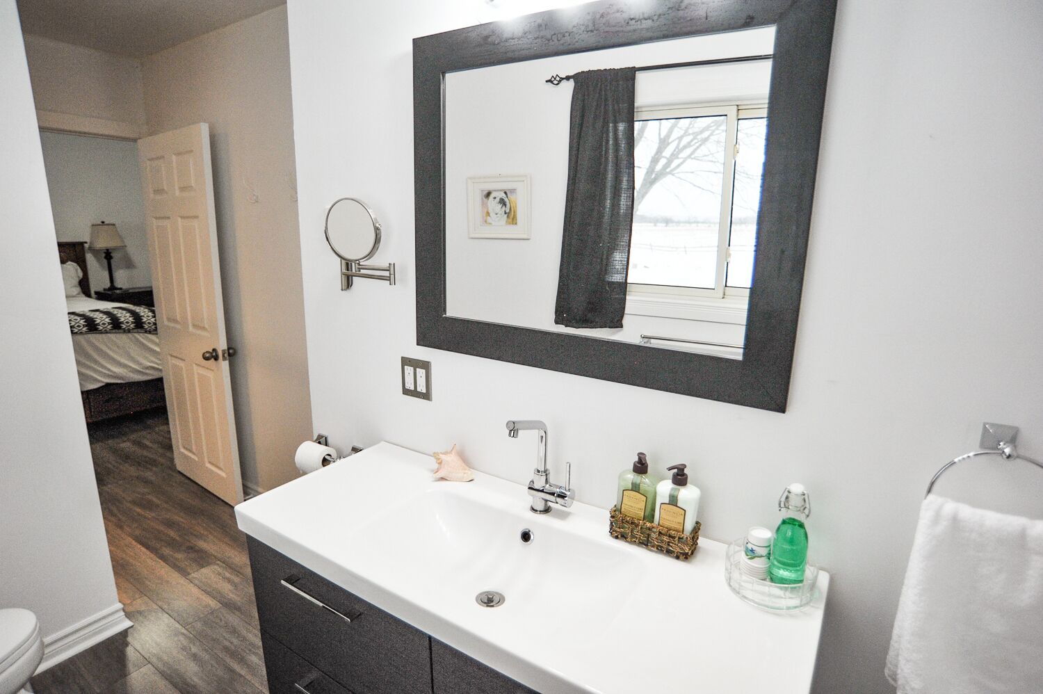 Une salle de bain avec un lavabo blanc, un miroir et des articles de toilette. Une porte donne sur une chambre à coucher équipée d’un lit et d’une lampe.