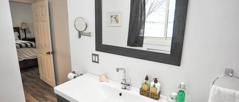 Une salle de bain avec un lavabo blanc, un miroir et des articles de toilette. Une porte donne sur une chambre à coucher équipée d’un lit et d’une lampe.