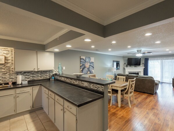 A modern kitchen with a central island, a dining area with a wooden table and chairs, a living room with a sofa and a TV, and a ceiling fan.