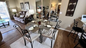 A dining area with a glass-top table, black chairs, and a vase with flowers. There is a sofa and a lamp in the background.