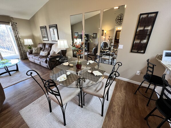 A dining area with a glass-top table, black chairs, and a vase with flowers. There is a sofa and a lamp in the background.