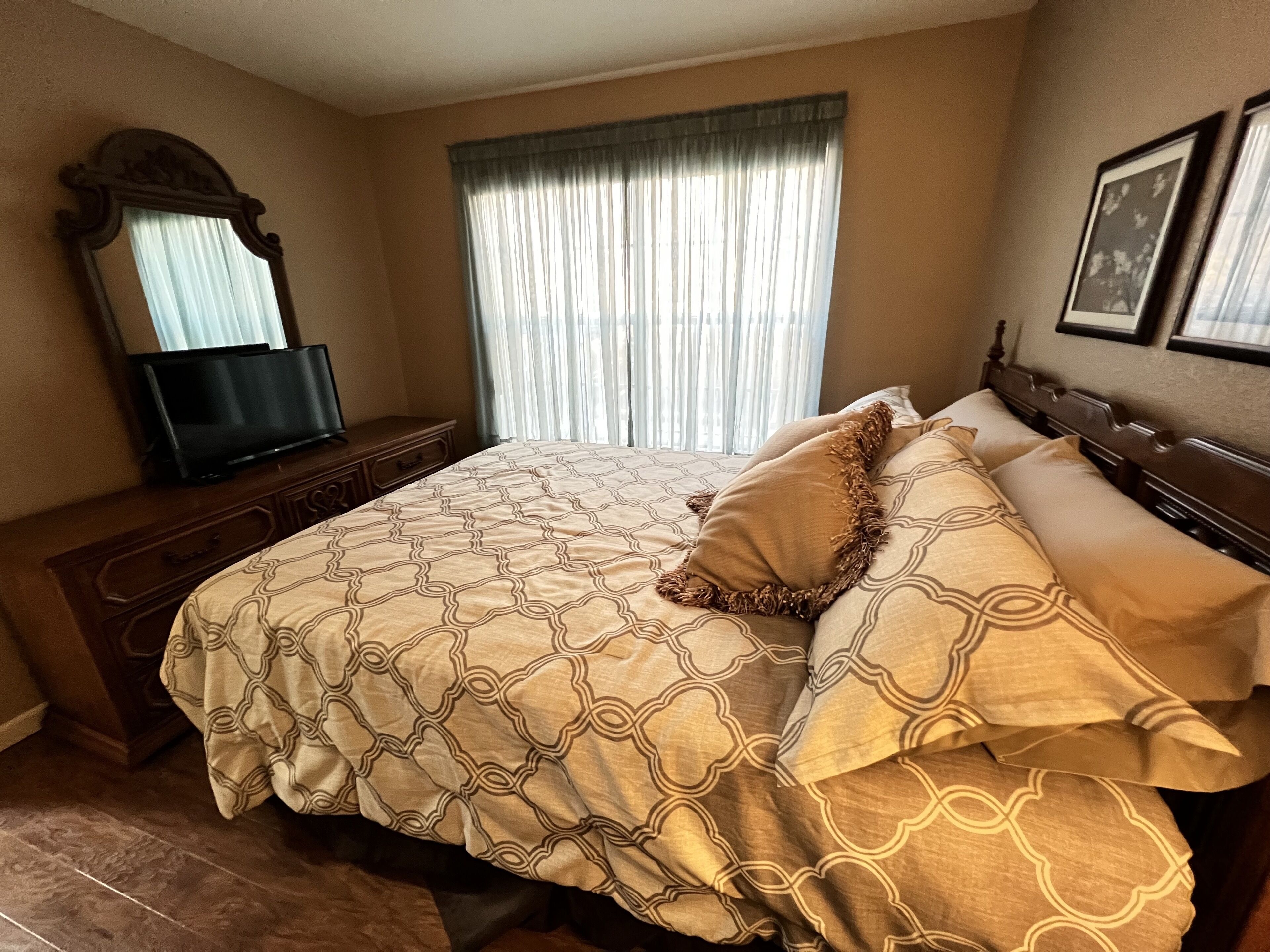 Un dormitorio con una cama, un televisor sobre un aparador de madera, una ventana con cortinas y cuadros enmarcados en la pared.