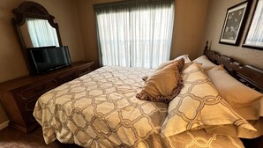 A bedroom with a bed, a television on a wooden dresser, a window with curtains, and framed pictures on the wall.