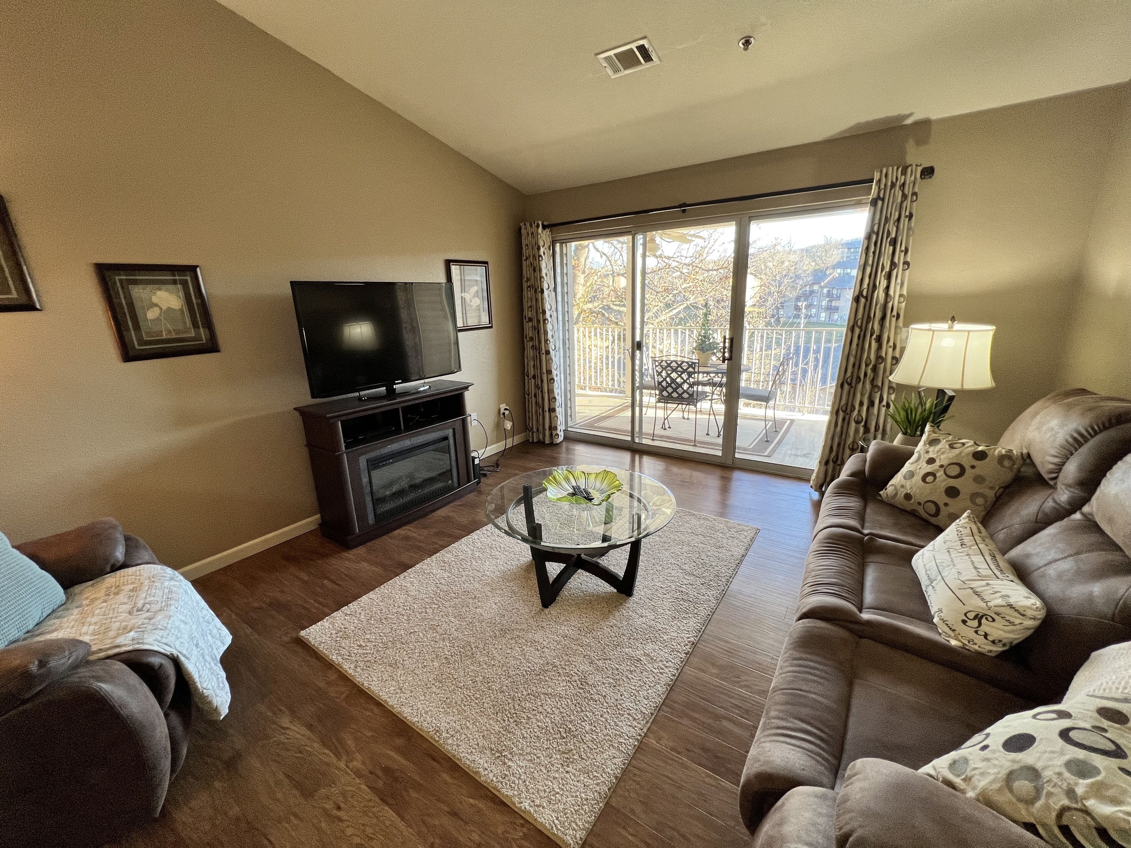 A living room with a fireplace, a flat-screen TV, a glass coffee table, and a sofa with patterned cushions.