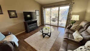 A living room with a fireplace, a flat-screen TV, a glass coffee table, and a sofa with patterned cushions.