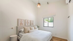 A bedroom with a large bed, a bedside table, a round waste basket, and a window with a view of greenery.