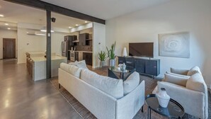 A modern living room with a sofa, a coffee table, and a TV. There is a kitchen in the background with cabinets and appliances.