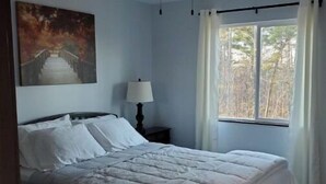 Ein Schlafzimmer mit Bett, Deckenventilator, Nachttisch mit Lampe, Vorhängen am Fenster und einem Bild an der Wand.