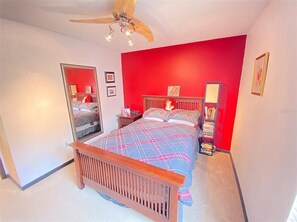 A bedroom with a wooden bed, a ceiling fan, a red accent wall, and a bedside table with a lamp.