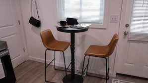 Ett litet rum med ett runt bord och två stolar, en svart handväska hängande på väggen och ett fönster med persienner.