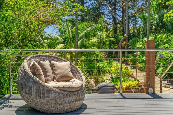 A wicker chair with cushions on a wooden deck, surrounded by lush greenery and a metal fence.