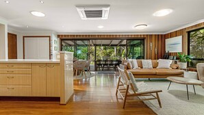 A spacious living area with a wooden floor, a brown leather sofa, a dining table with chairs, and a kitchen area with wooden cabinets and a white countertop.