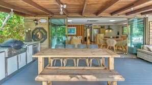 An outdoor kitchen area with a wooden table and bar stools, a grill, and a glass door leading to a dining area with a sofa and chairs.
