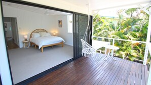 Ein Schlafzimmer mit Bett, Nachttisch und Lampe, verbunden mit einem Balkon mit Hängesessel und Tisch, mit Blick auf einen tropischen Garten.