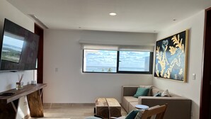 Una sala moderna con un televisor grande, una mesa de centro de madera, un sofá y una silla. Se ve una ventana con vistas al océano y un cuadro en la pared.