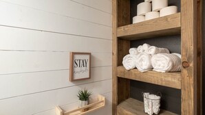 A wooden shelf with rolled towels and a framed sign saying "STAY awhile" on a white wooden wall. - Studied Perfection (Spearfish)