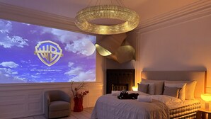 Un dormitorio con cama, una silla, un televisor que muestra el logotipo de Warner Bros. Television y una lámpara decorativa en el techo.