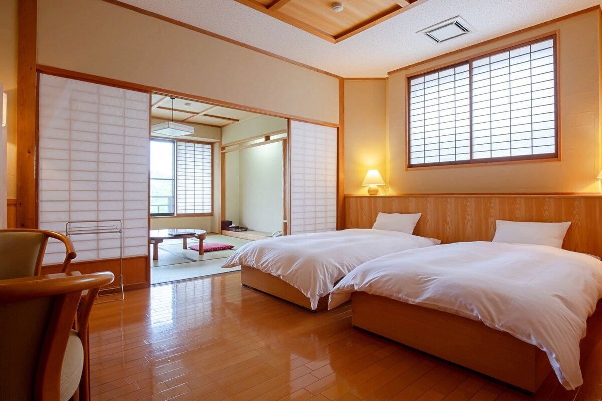 Habitación de hotel con piso de madera, dos camas, una silla y una puerta corredera que da a un baño.