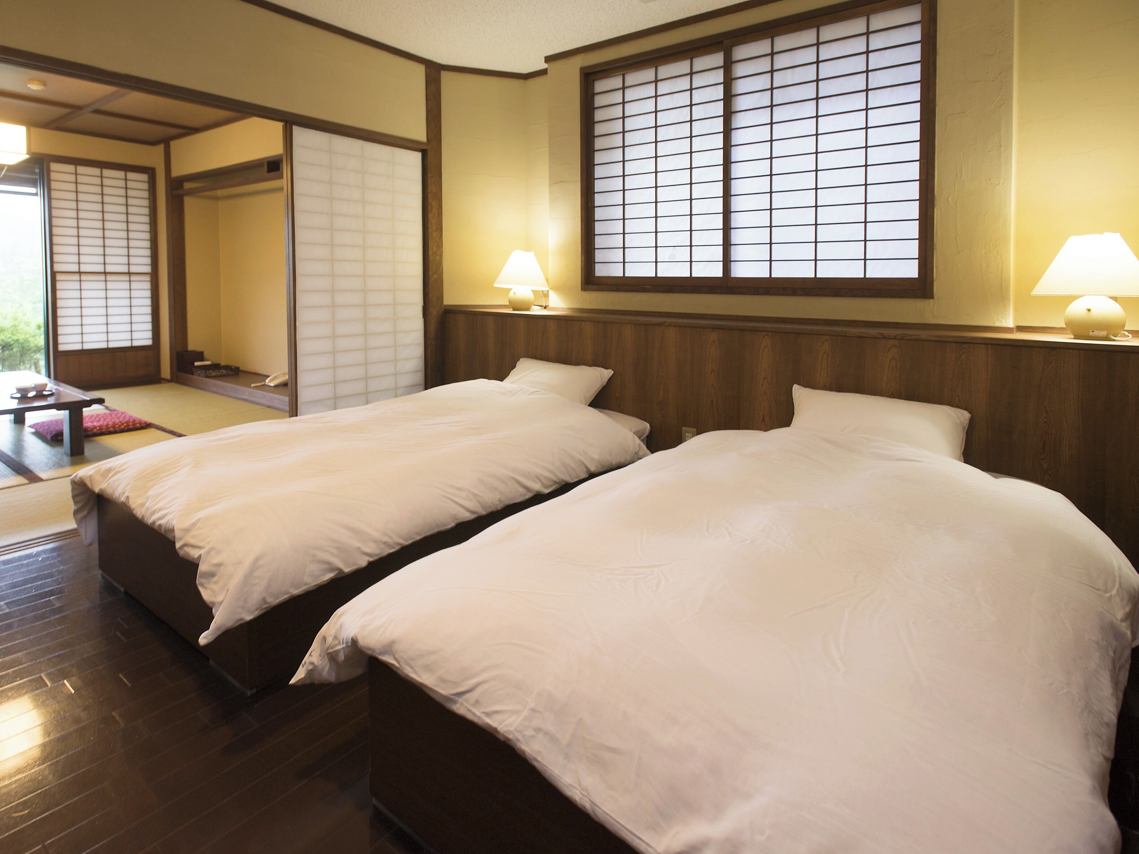 A hotel room with two beds, a wooden floor, a sliding door, and a window with a view.