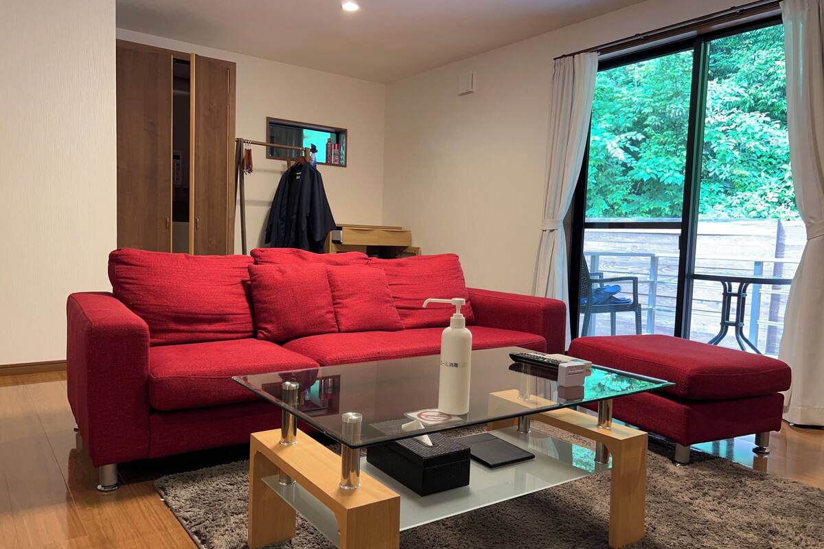 Un canapé rouge, une table basse en verre et une vue sur la verdure par une grande fenêtre.