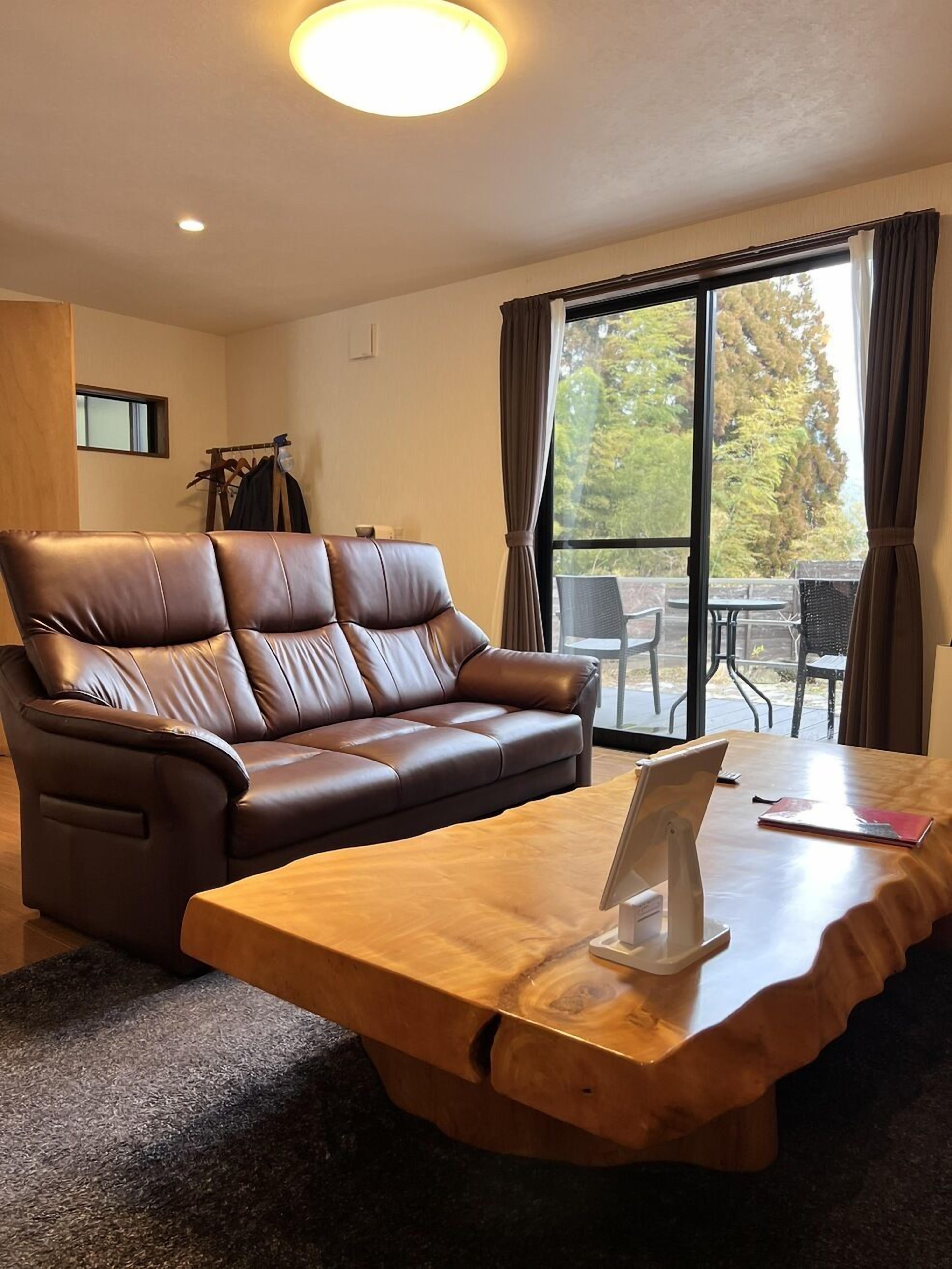 Ein Zimmer mit einem braunen Ledersofa, einem Holztisch und Blick auf Gartenmöbel durch ein großes Fenster.