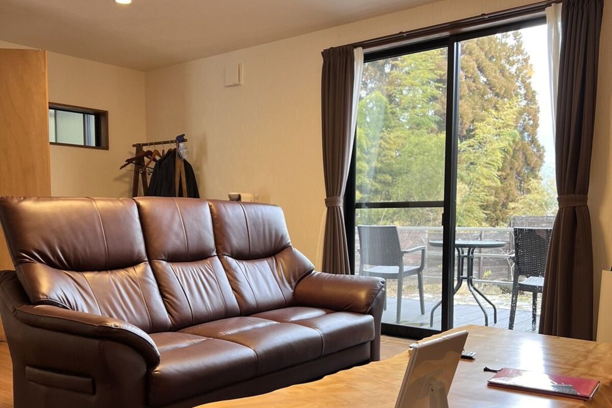 A room with a brown leather sofa, a wooden coffee table, and a view of outdoor furniture through a large window.