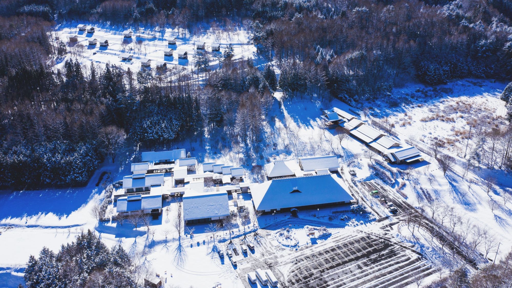 Eine Vogelperspektive zeigt eine verschneite Landschaft mit mehreren Gebäuden, darunter ein großes Hauptgebäude mit geneigtem Dach, umgeben von kleineren Gebäuden und dichtem Wald.