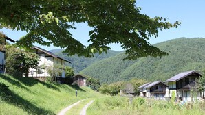 A rural setting with a dirt path leading to a row of traditional houses with sloped roofs, surrounded by lush greenery and hills in the background. - Green Village Yokokawa Kayabuki No Yakata (Tatsuno)