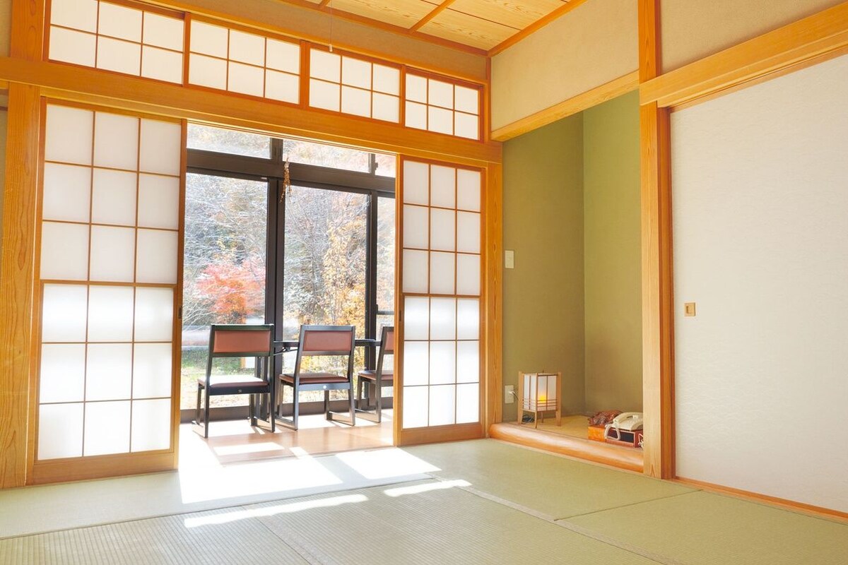 A traditional Japanese room with tatami flooring, sliding doors, and a view of autumn foliage.