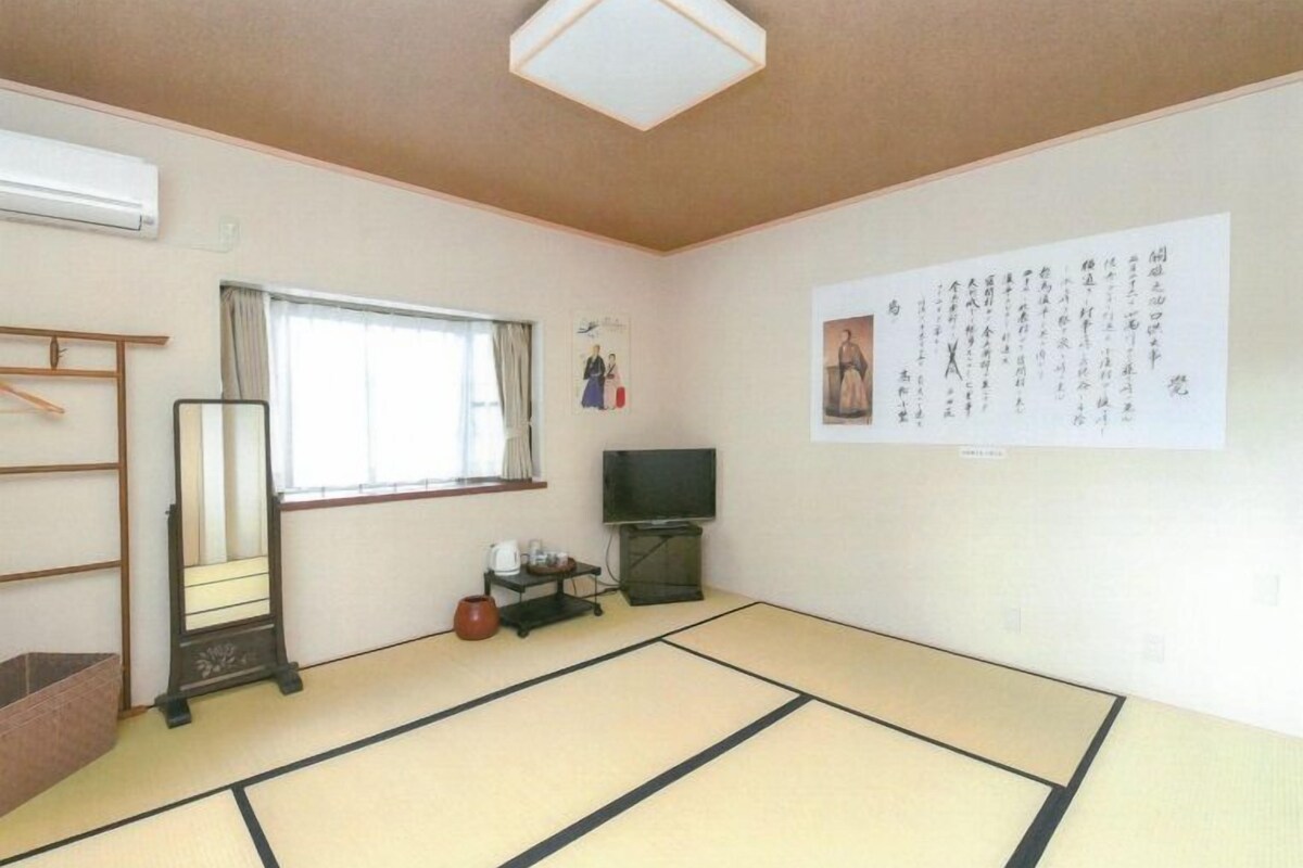 A room with tatami flooring, a television, a shelf with items, a window with curtains, and a wall with calligraphy.